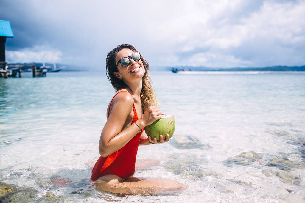 Πορτρέτο του αστείο Καυκάσιο κορίτσι σε μοντέρνα γυαλιά ηλίου δροσιστικό το καλοκαίρι στη θάλασσα κρατώντας καρύδα φρούτων και χαμογελώντας στην κάμερα, χαρούμενη γυναίκα με φυσικό ποτό ποζάρουν στην παραλία των τροπικών Κέιμαν - Φωτογραφία, εικόνα