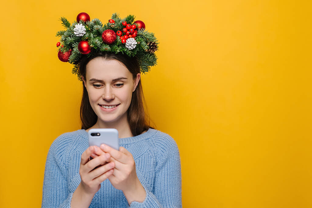 Задоволена молода жінка друкує текстове повідомлення на мобільному телефоні, насолоджується онлайн-спілкуванням, друкує зворотній зв'язок, носить різдвяний вінок і зимовий светр, ізольований на жовтому тлі. Концепція Різдва
 - Фото, зображення