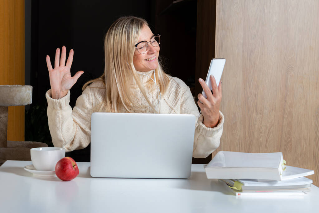 Frau mit Kopfhörer mit Handy während der Arbeit mit Laptop in der heimischen Küche, Online-Lebensstil - Foto, Bild