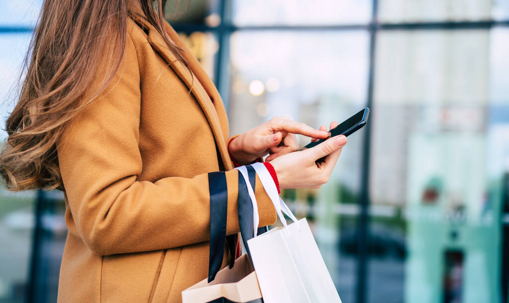 Όμορφη μοντέρνα νεαρή γυναίκα με πολλά πολύχρωμα τσάντες ψώνια σε καλή διάθεση με έξυπνο τηλέφωνο και πιστωτική κάρτα, ενώ το περπάτημα στο εμπορικό κέντρο κατά τη διάρκεια της μαύρης Παρασκευής - Φωτογραφία, εικόνα