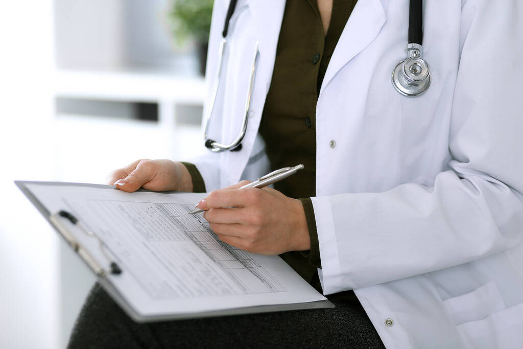 Woman doctor writing something at clipboard while sitting at the chair, close-up. Therapist at work filling up medication history records. Medicine and healthcare concept - Photo, Image