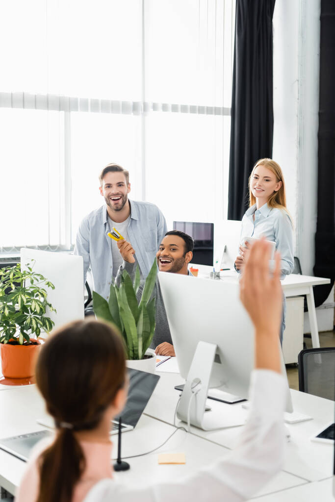 Uśmiechnięci wielonarodowi biznesmeni patrzący na kolegę z podniesioną ręką na niewyraźnym pierwszym planie w biurze  - Zdjęcie, obraz