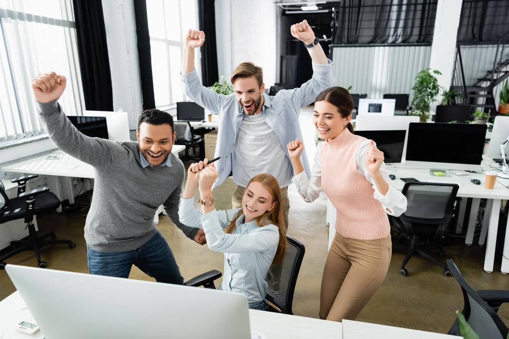 Positive multiethnic businesspeople showing yeah gesture near computer on blurred foreground in office  - Photo, Image