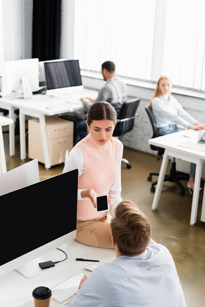 Empresária segurando smartphone com tela em branco perto de colega e computador no escritório  - Foto, Imagem