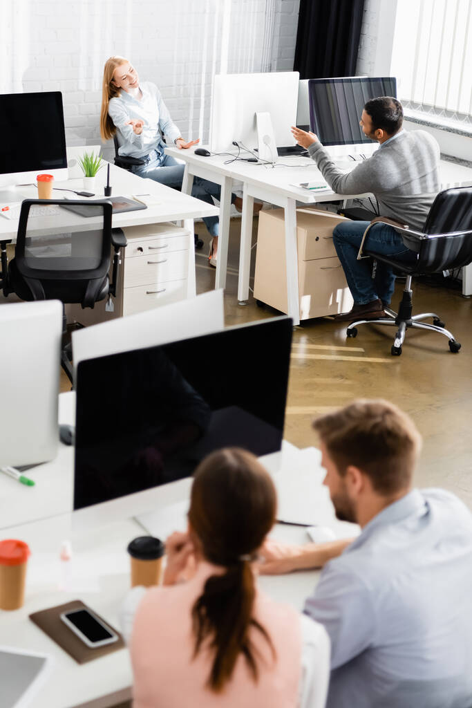 Улыбающиеся мультиэтнические предприниматели разговаривают за компьютерами в офисе  - Фото, изображение
