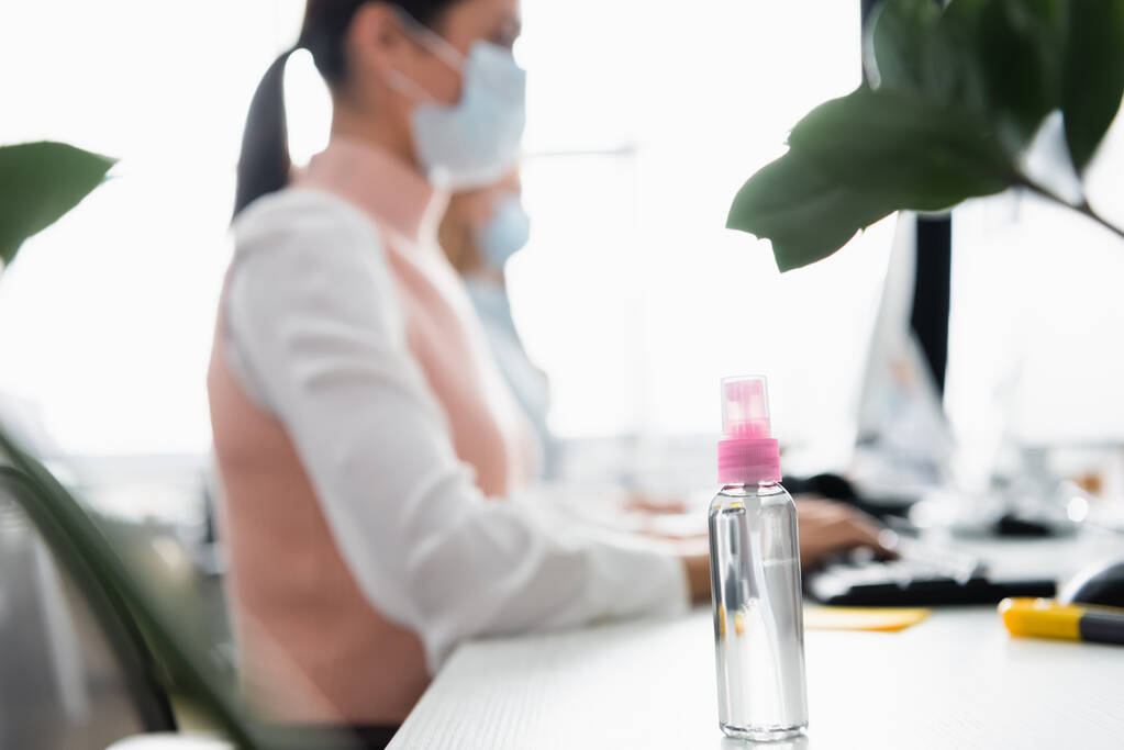 Средство для дезинфекции рук на столе рядом с предпринимательницей в медицинской маске, работающей на размытом фоне  - Фото, изображение