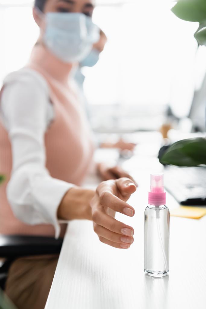Bizneswoman w masce medycznej ciągnąc rękę do środka dezynfekującego na stole na rozmytym tle  - Zdjęcie, obraz