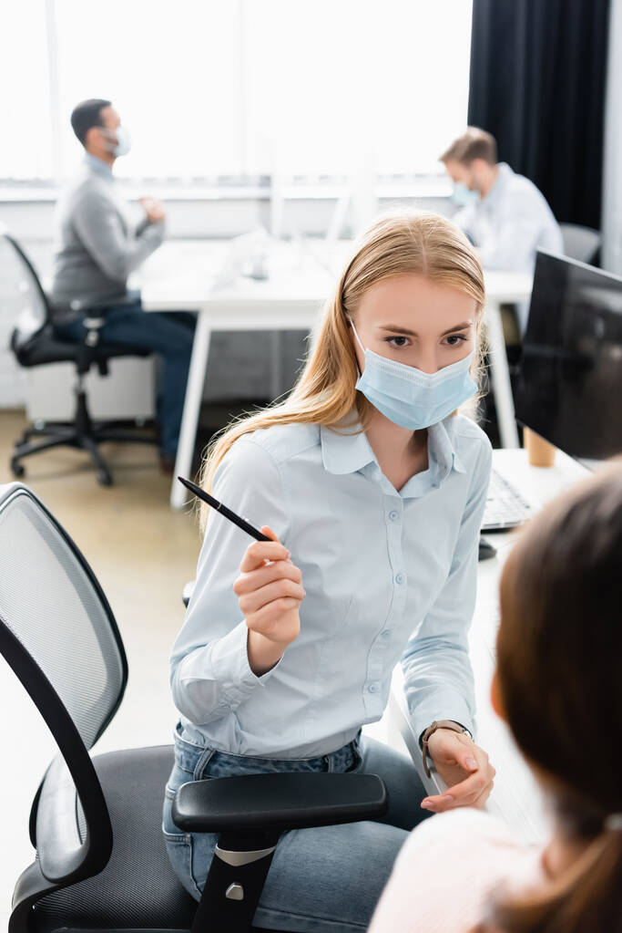 Empresaria en máscara médica sosteniendo pluma cerca de colega en primer plano borrosa  - Foto, imagen