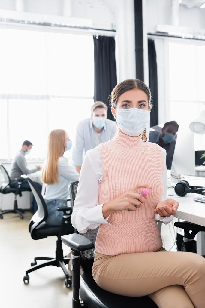 Zakenvrouw in medisch masker met behulp van de hand sanitizer terwijl collega 's werken op wazig achtergrond  - Foto, afbeelding