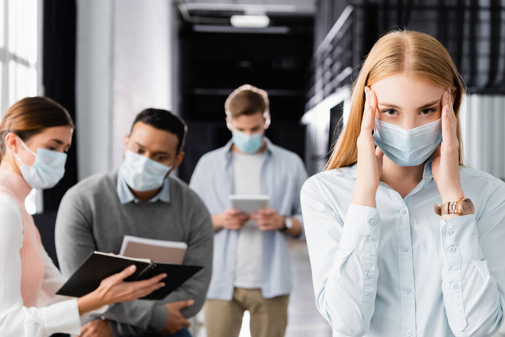 Diseased businesswoman in medical mask touching near while multiethnic colleagues working at blurred background  - Photo, Image