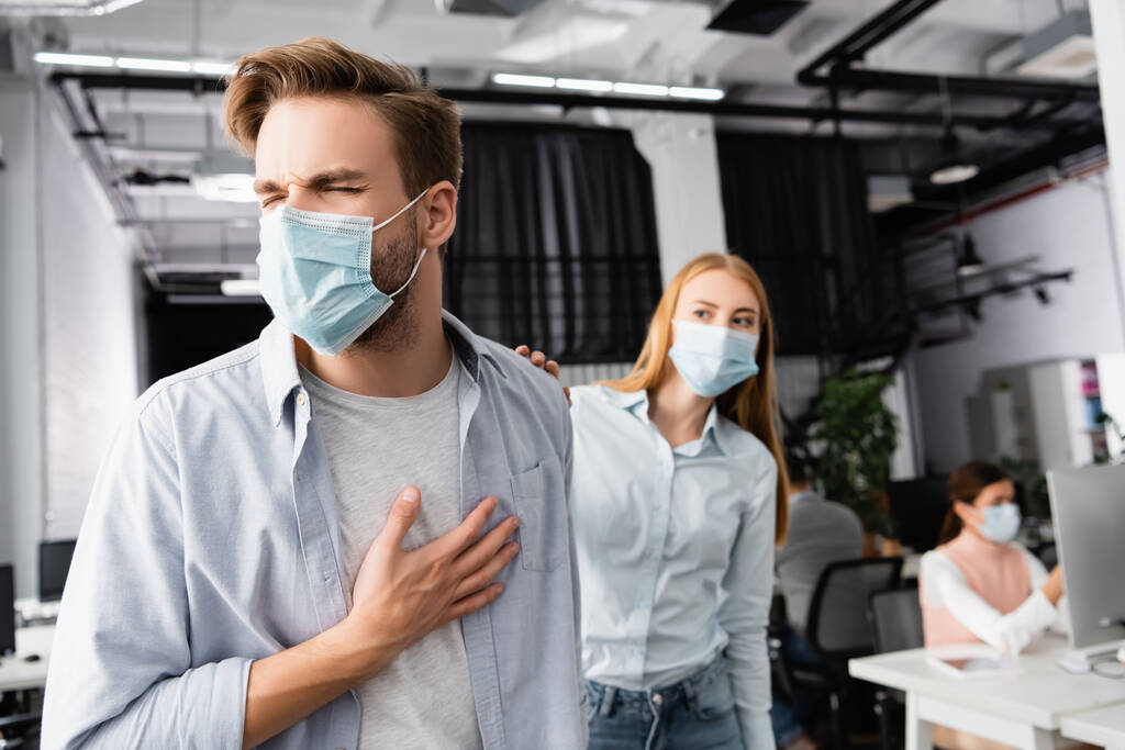 Empresario enfermo en máscara médica tocando el pecho cerca de colega en fondo borroso en la oficina  - Foto, imagen