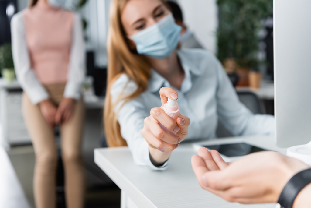 Sağlık maskeli bir iş kadını ofisteki bulanık ön planda meslektaşının eline el dezenfektanı sıkıyor.  - Fotoğraf, Görsel