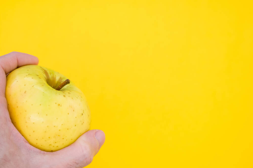 黄色の背景に単離された黄色のリンゴを保持する手のクローズアップショット - 写真・画像