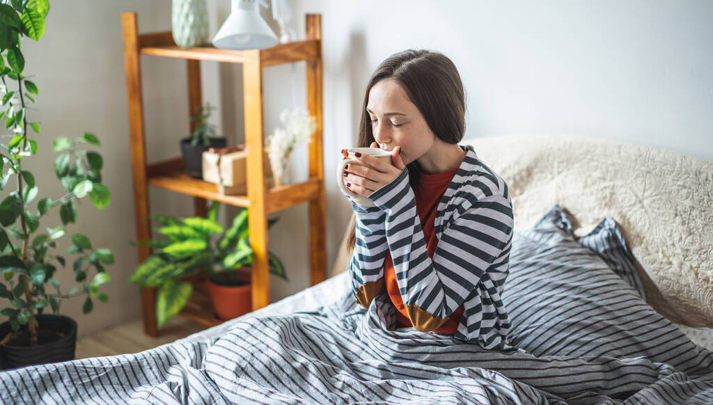 Fiatal, gyönyörű lány pizsamában ül az ágyon egy napos reggelen és kávét iszik. Fogalom a hangulat egy jó nap. - Fotó, kép