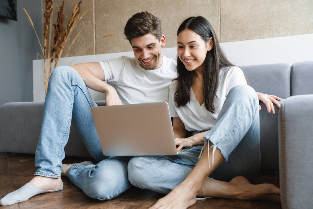 Zdjęcie wesołej młodej kochającej się pary w domu na piętrze przy sofie przy użyciu laptopa - Zdjęcie, obraz
