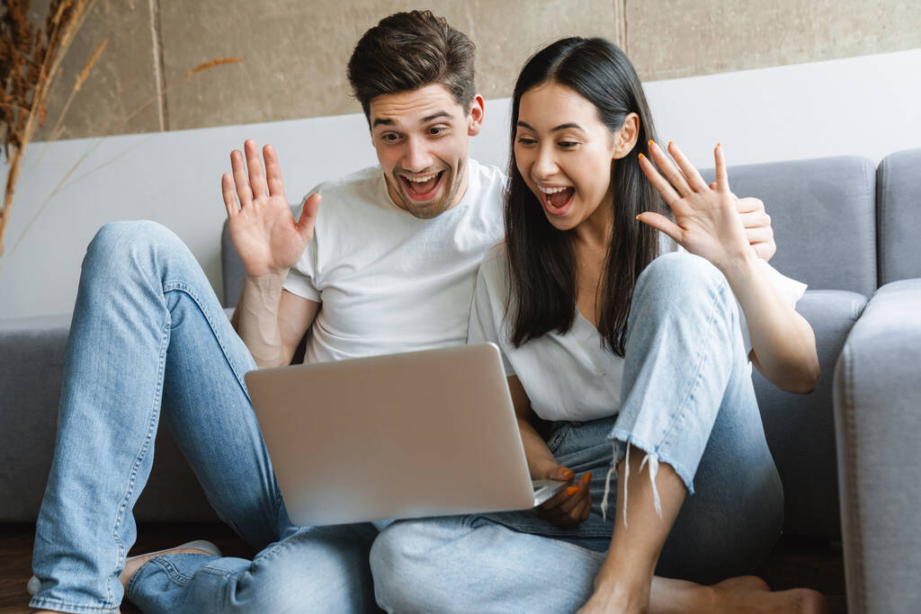 ソファーの近くの床の上の自宅で肯定的な若い愛のカップルの写真ビデオコールによってラップトップコンピュータと手を振っ - 写真・画像