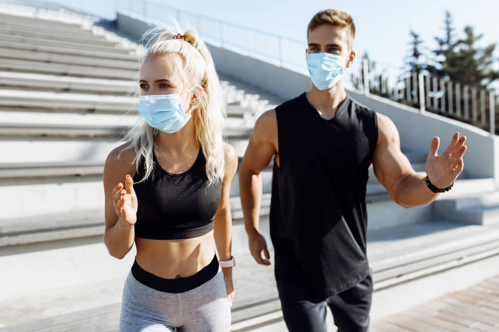 Genç spor çifti erkek ve kadın, yüzlerinde tıbbi koruyucu maskeler ve spor kıyafetlerle, şehirde cadde boyunca birlikte koşuyorlar, karantina, koronavirüs. - Fotoğraf, Görsel