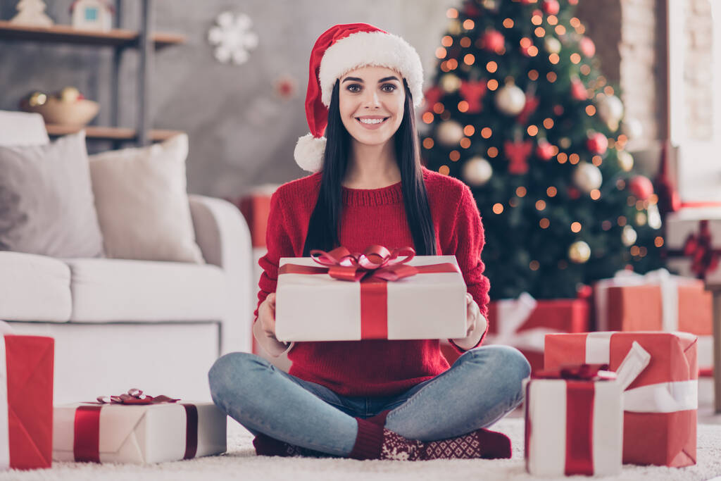 Photo of pretty young girl sit floor hold big present box wear santa headwear red sweater jeans socks in decorated living room indoors - Photo, Image