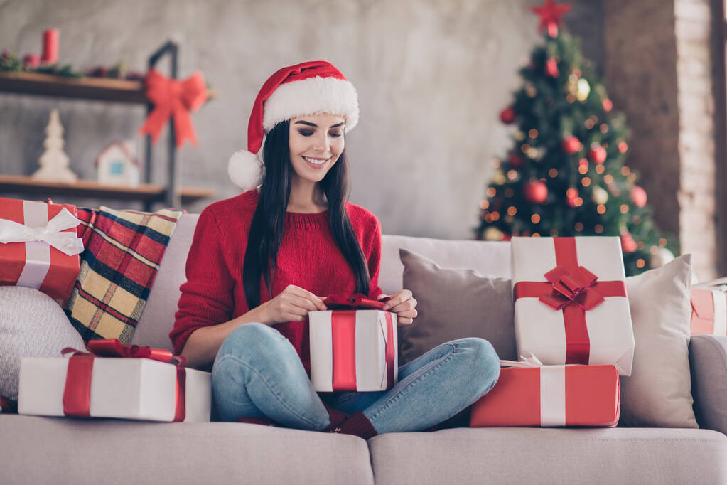 Foto di bella ragazza seduta divano tenere scartando giftbox molti regali indossare Santa cap jeans maglione rosso in salotto decorato all'interno - Foto, immagini