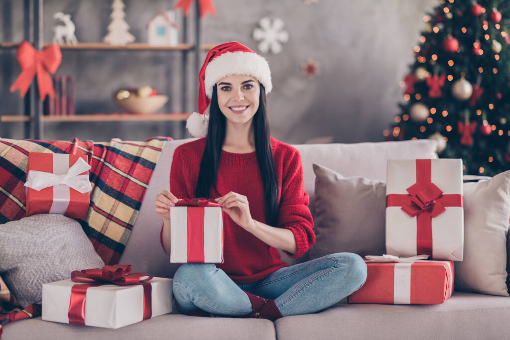 Kuva söpö nuori nainen istua sohvalla pitää lahjapaketti keula monia lahjoja kulumista Santa päähineet punainen villapaita farkut sisustettu olohuoneessa sisätiloissa - Valokuva, kuva