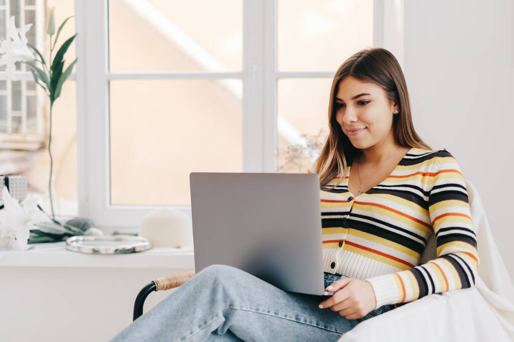Atrakcyjna młoda biała kobieta używa laptopa, siedząc na fotelu w jasnym salonie. Koncepcja pracy na odległość - Zdjęcie, obraz
