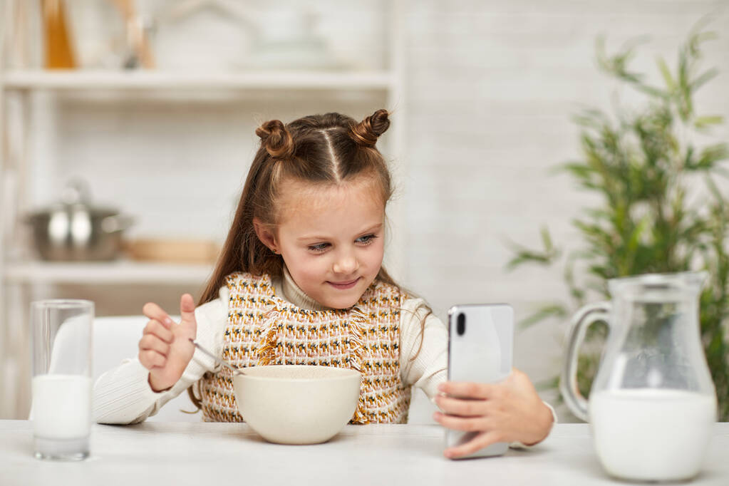 klein meisje eten ontbijt en kijken naar de smartphone - Foto, afbeelding