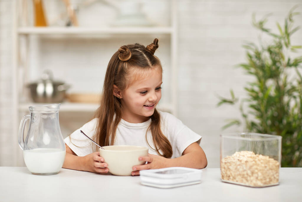 dziewczynka jedząca śniadanie: płatki zbożowe z mlekiem w kuchni. - Zdjęcie, obraz