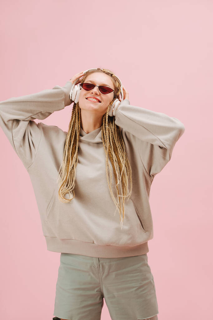 Radosna młoda kobieta w wąskich okularach przeciwsłonecznych ze stylowymi blond afro warkoczami słuchająca muzyki, przyciskająca słuchawki do głowy. Na różowym tle. - Zdjęcie, obraz