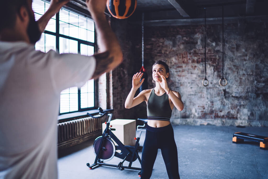 Позитивна біла жінка в активному носінні кидає м'яч до свого тренера під час тренування для схуднення, жінка схуднення зосереджена на фізичних вправах, щоб схуднути і зберегти ідеальну форму тіла
 - Фото, зображення