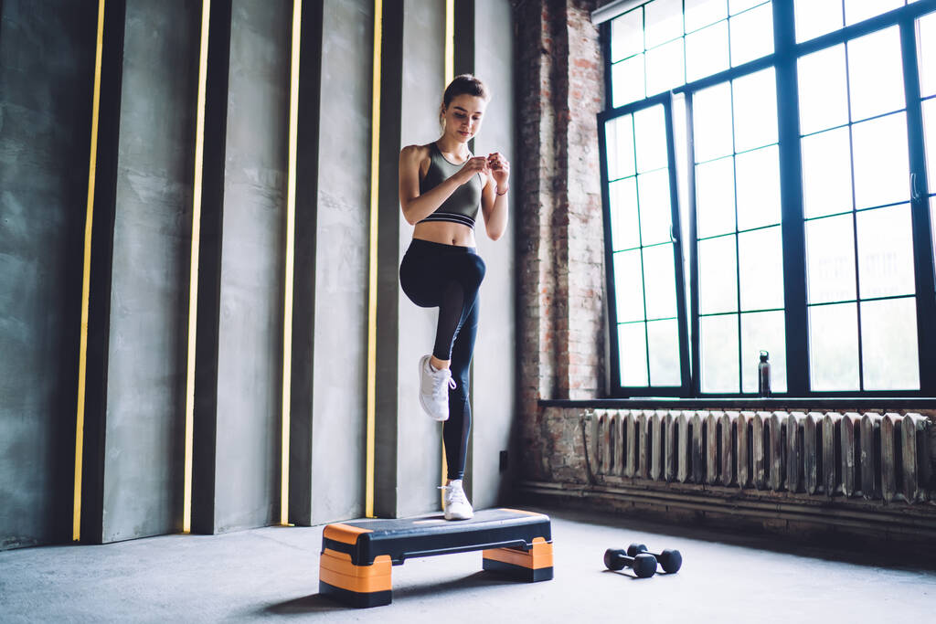 Полное тело стройная сконцентрированная женщина в спортивной одежде делает высокие колено марш упражнения на ступенчатой платформе в легком просторном тренажерном зале - Фото, изображение