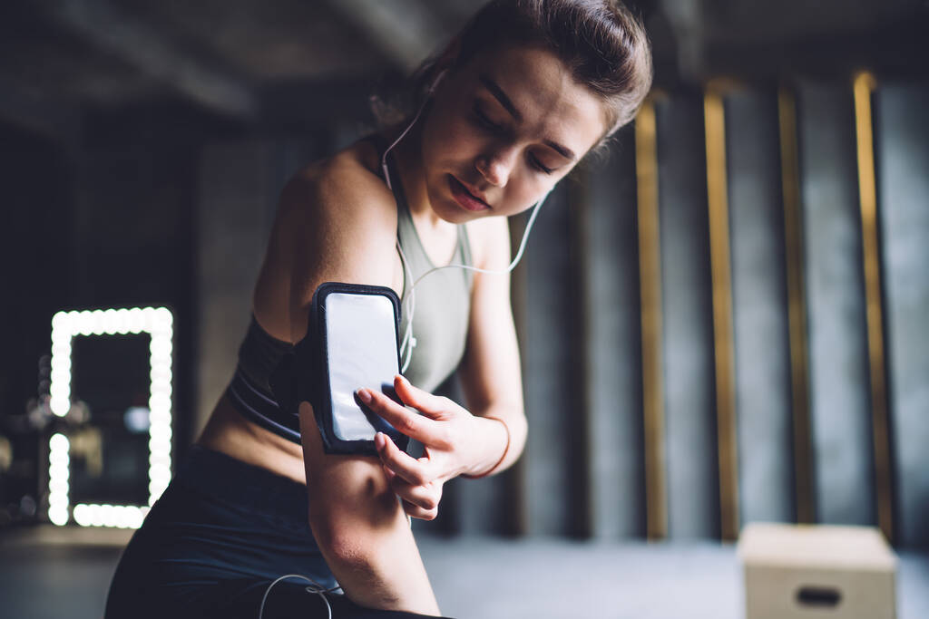 Atleta femenina delgada en ropa deportiva de pie en el gimnasio y el teléfono inteligente de navegación en el brazalete mientras escucha música en los auriculares durante el entrenamiento - Foto, imagen