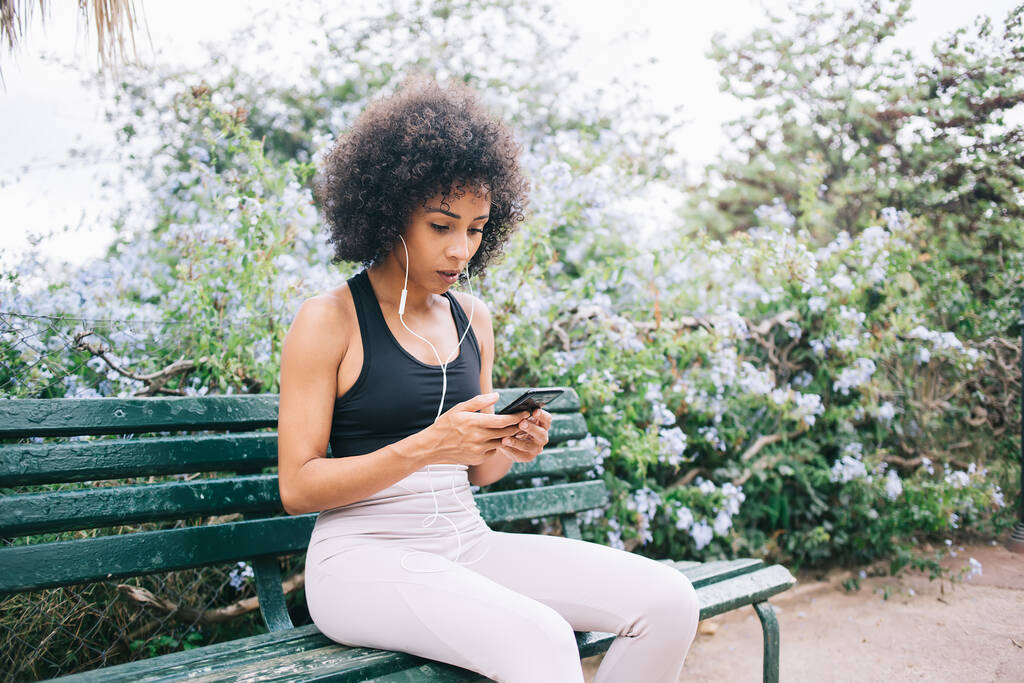 Atleta femenina concentrada en ropa deportiva mensajería en el teléfono móvil y escuchar audio con auriculares mientras descansa en el banco en el parque verde - Foto, Imagen