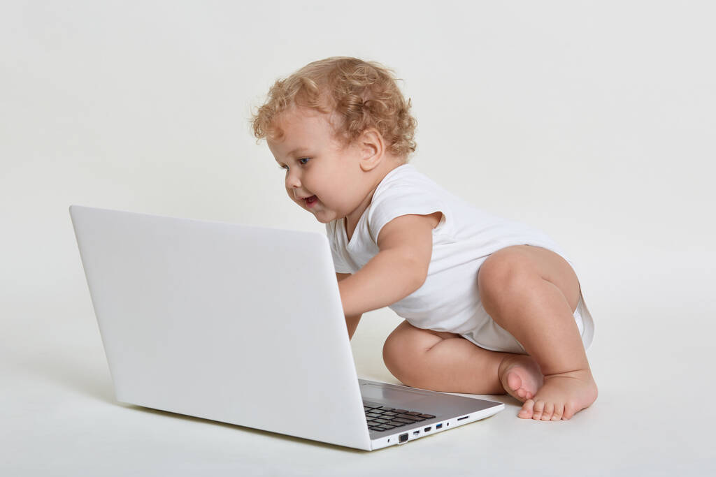 Entzückendes Baby, das auf dem Boden sitzt und mit aufgeregtem Gesichtsausdruck auf den Laptop-Bildschirm blickt, Säugling im weißen Ganzkörperanzug, blondes gelocktes Haar, Tastatur mit der Hand anfassen. - Foto, Bild