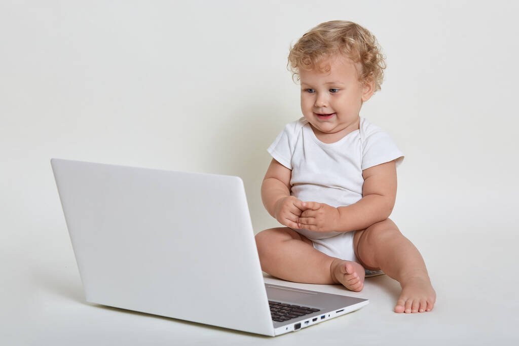 Charming blond curly haired infant wearing bodysuit, sitting barefoot on floor and looking directly at screen of lap top, barefoot toddler against white background. - Photo, Image