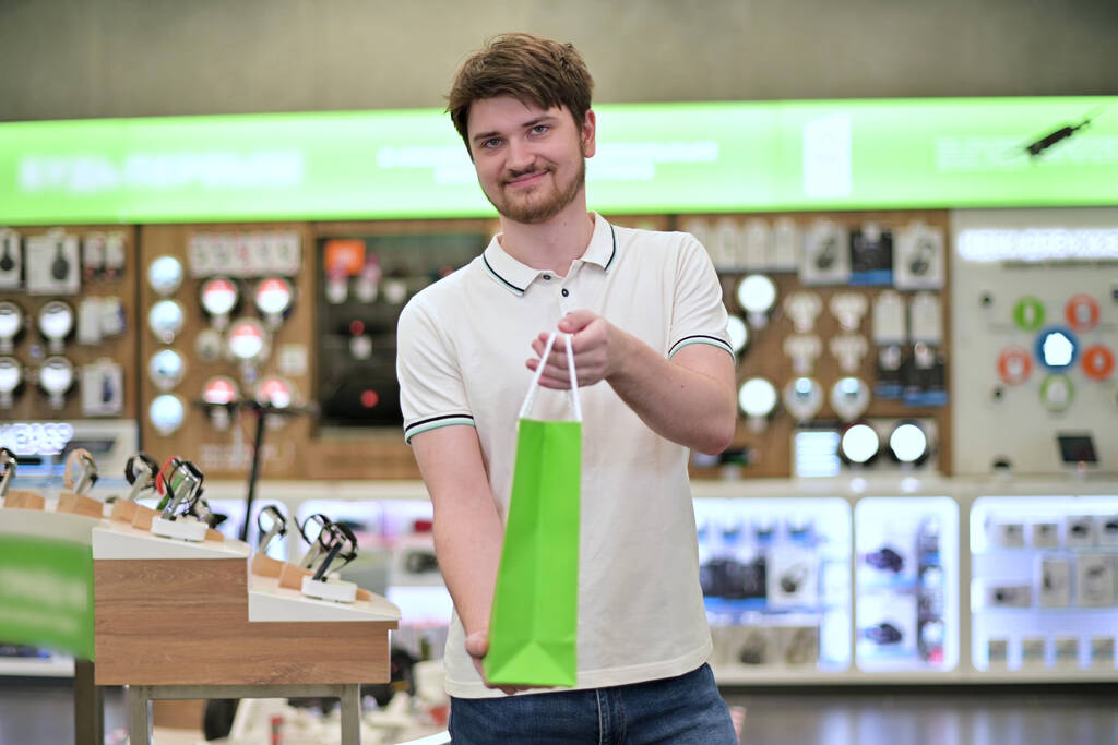 Joven vendedor en la tienda de electrónica pone mercancías en la bolsa de papel y sostiene a la cámara, sonriendo lindo - Foto, Imagen