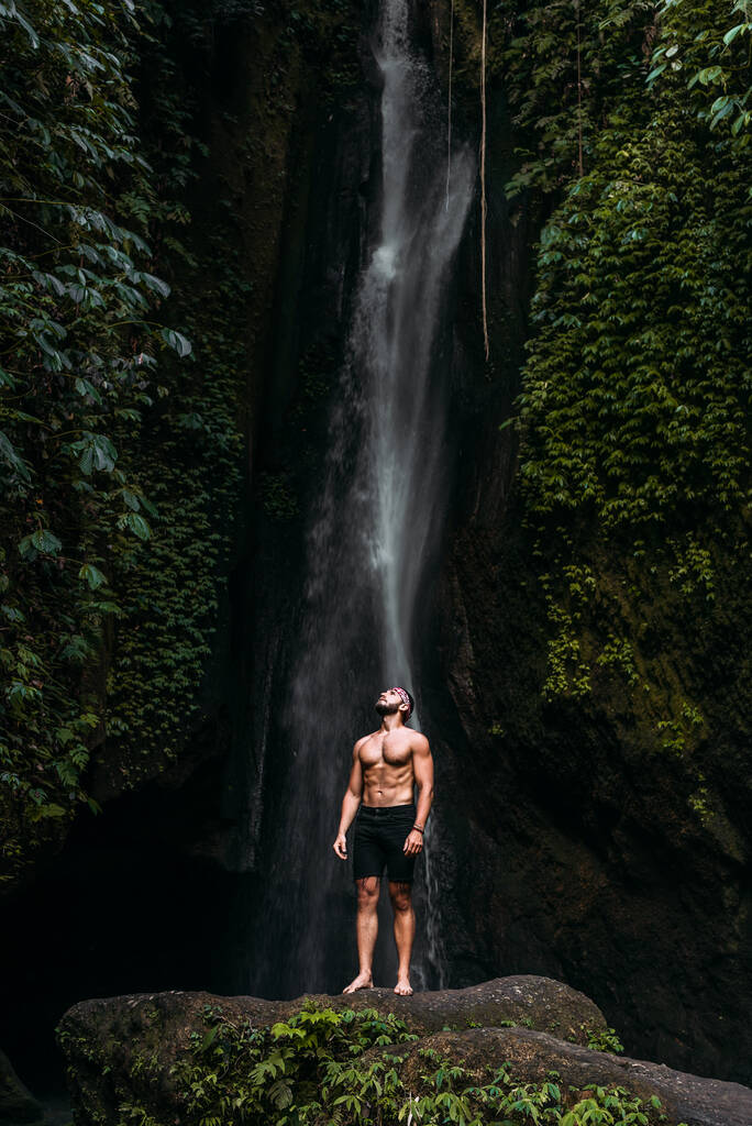 滝に運動用の建物の男。男は世界を旅する。滝の男。バリ島インドネシアへの旅行。絵のように美しい場所を旅する人。一人旅。スペースのコピー - 写真・画像