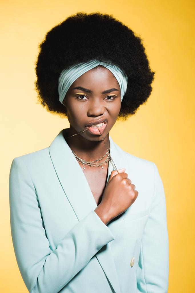 アフリカ系アメリカ人の若い女性の青いスタイリッシュな服とネックレスの口の中に黄色に隔離された - 写真・画像
