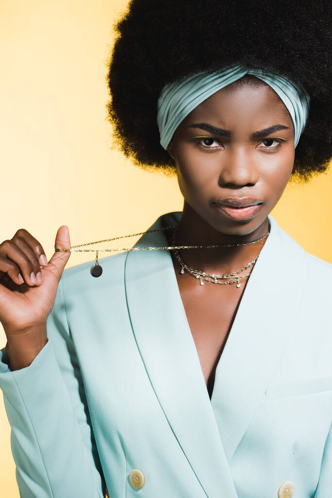 seria afroamericana giovane donna in abito elegante blu mostrando collana isolata sul giallo - Foto, immagini