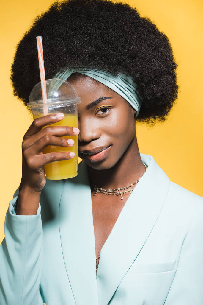 africká americká mladá žena v modrém stylovém oblečení s pomerančovou šťávou izolované na žluté  - Fotografie, Obrázek