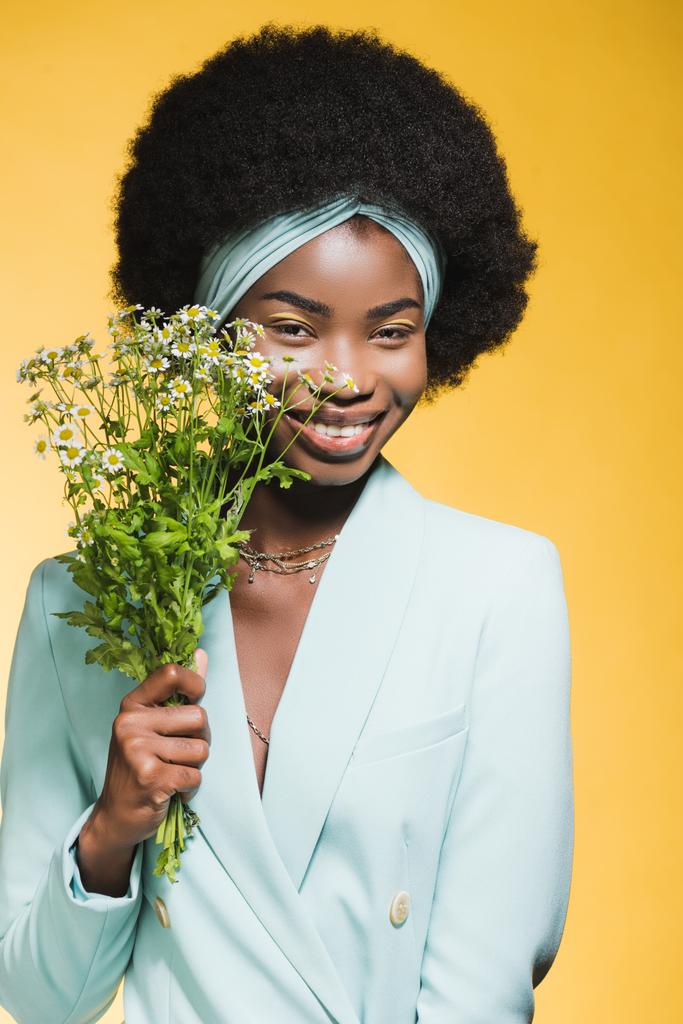 usmívající se africká americká mladá žena v modrém stylovém oblečení s heřmánkovou kyticí izolované na žluté  - Fotografie, Obrázek