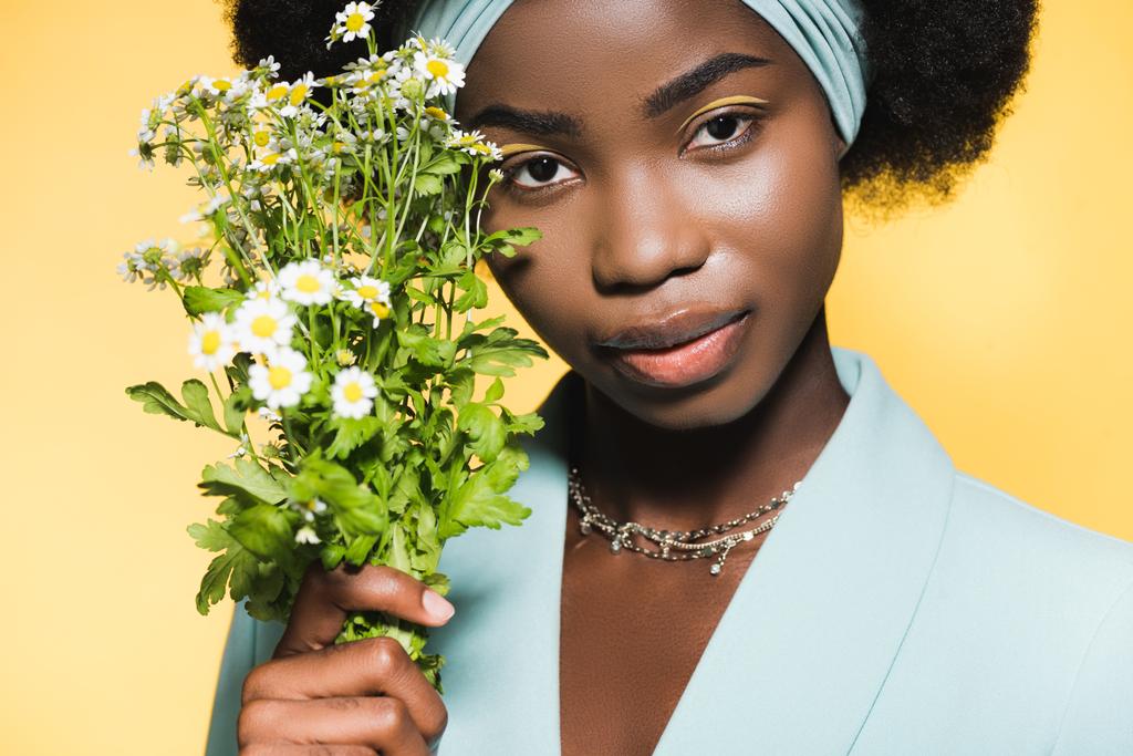 african american young woman in blue stylish outfit with chamomile bouquet isolated on yellow  - Photo, Image