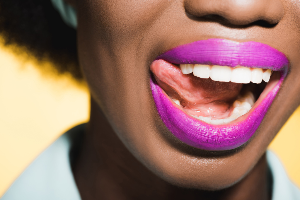 bijgesneden weergave van Afrikaanse Amerikaanse jonge vrouw met paarse lippen tonen tong geïsoleerd op geel  - Foto, afbeelding