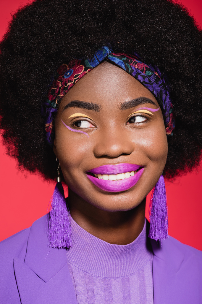 χαμογελαστή Αφροαμερικανή νεαρή γυναίκα με μοβ στυλάτο ντύσιμο απομονωμένη στο κόκκινο - Φωτογραφία, εικόνα