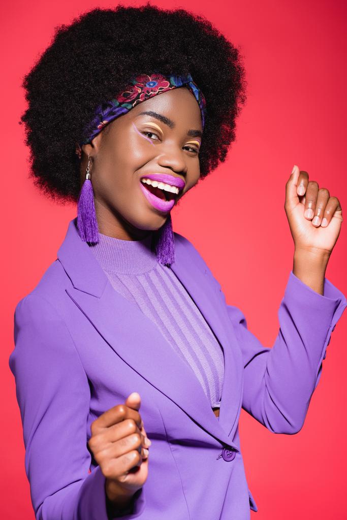 happy african american young woman in purple stylish outfit isolated on red - Photo, Image
