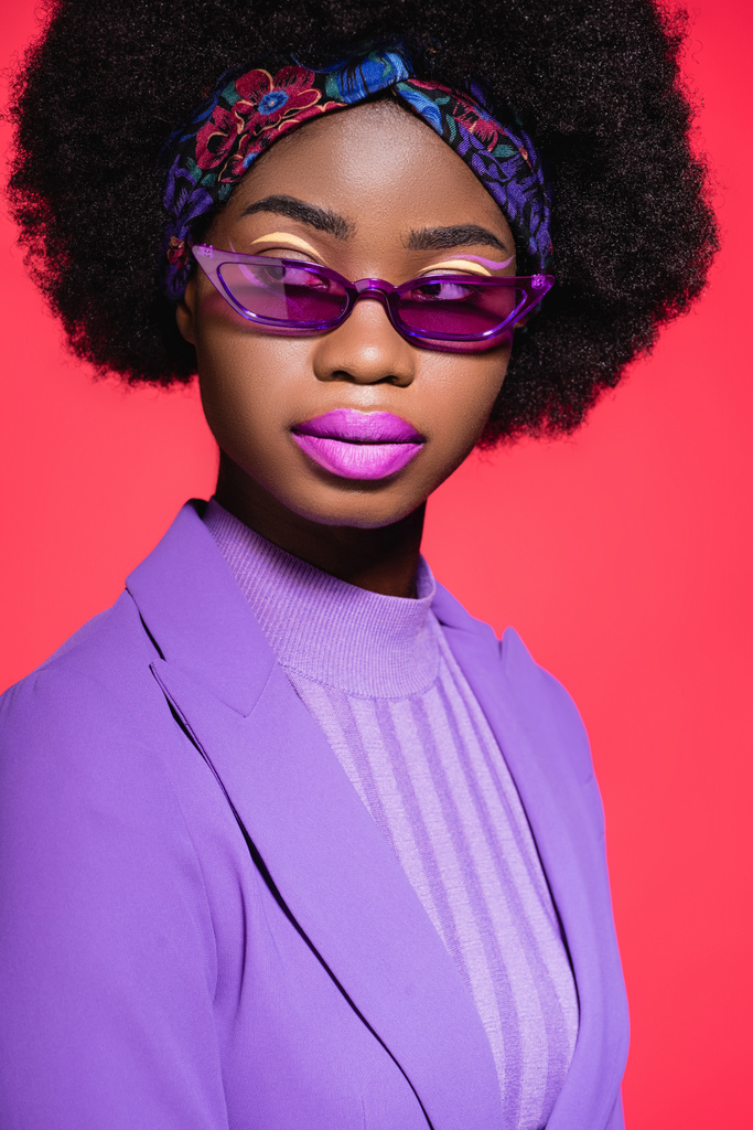Африканская американская молодая женщина в фиолетовом стильном наряде и солнечных очках, изолированных на красном - Фото, изображение