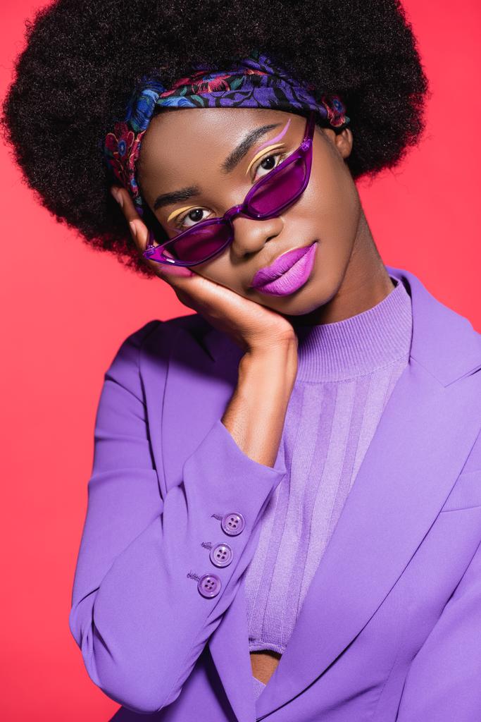 Африканская американская молодая женщина в фиолетовом стильном наряде и солнечных очках, изолированных на красном - Фото, изображение