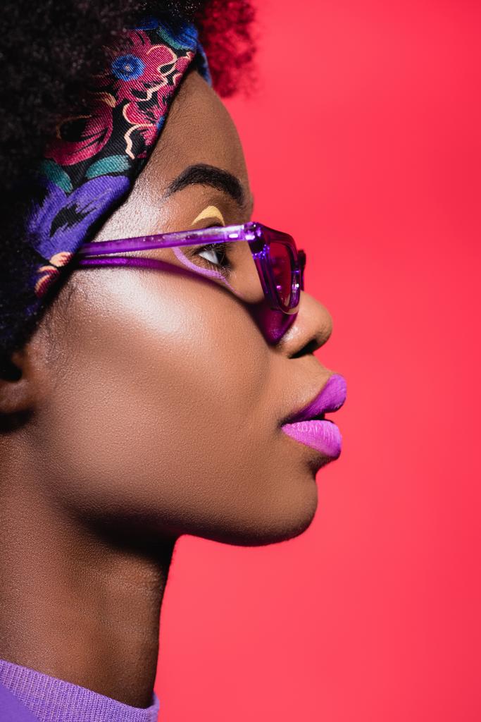 profile of african american young woman in purple stylish outfit and sunglasses isolated on red - Photo, Image