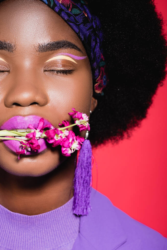 обрізаний вид афроамериканської молодої жінки в фіолетовому стильному вбранні з квіткою в роті ізольовано на червоному
 - Фото, зображення