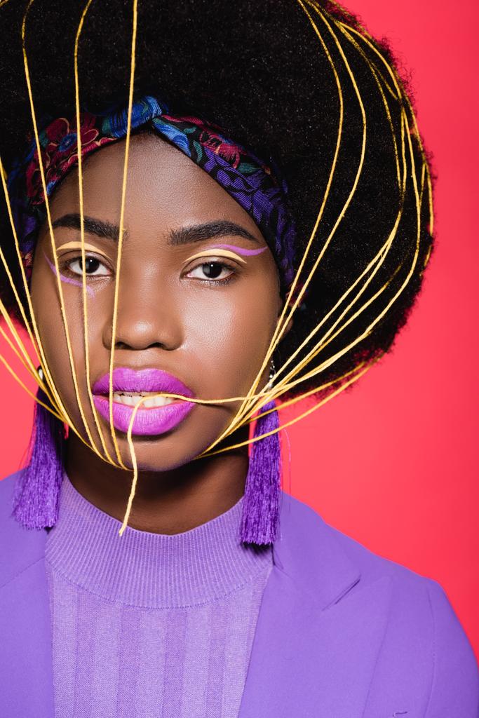 african american young woman in purple stylish outfit with yellow strings on face isolated on red - Photo, Image