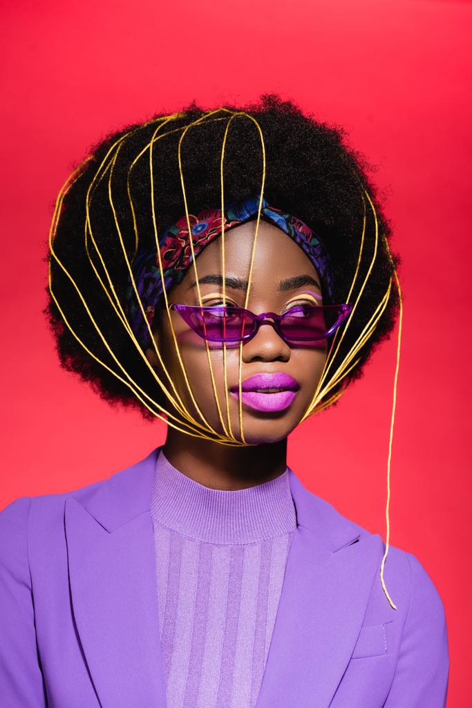 africký americký mladý žena v fialové stylové oblečení a sluneční brýle se žlutými strunami na obličeji izolované na červené - Fotografie, Obrázek